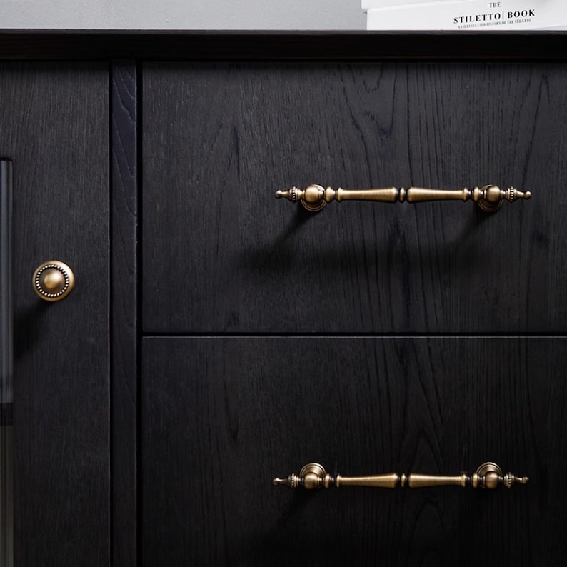 Peut inclure: Gros plan sur une armoire en bois noir avec deux tiroirs et une porte. L'armoire est orn&eacute;e de quincaillerie en laiton antique, notamment deux poign&eacute;es de tiroir et un bouton de porte rond. Le grain du bois est visible, ajoutant de la texture &agrave; la surface sombre.