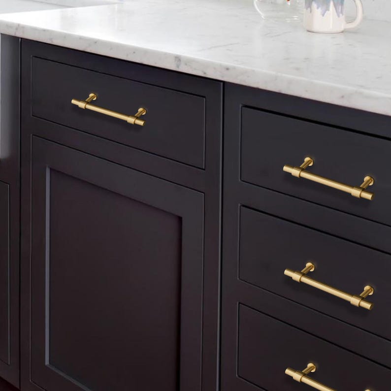 K&ouml;nnte beinhalten: Nahaufnahme eines K&uuml;chenschranks mit schwarz lackierten T&uuml;ren und Schubladen. Goldfarbene Stangengriffe sind an den Schubladen angebracht. Die Arbeitsplatte ist aus wei&szlig;em Marmor. Der Schrank ist Teil einer K&uuml;chenzeile.