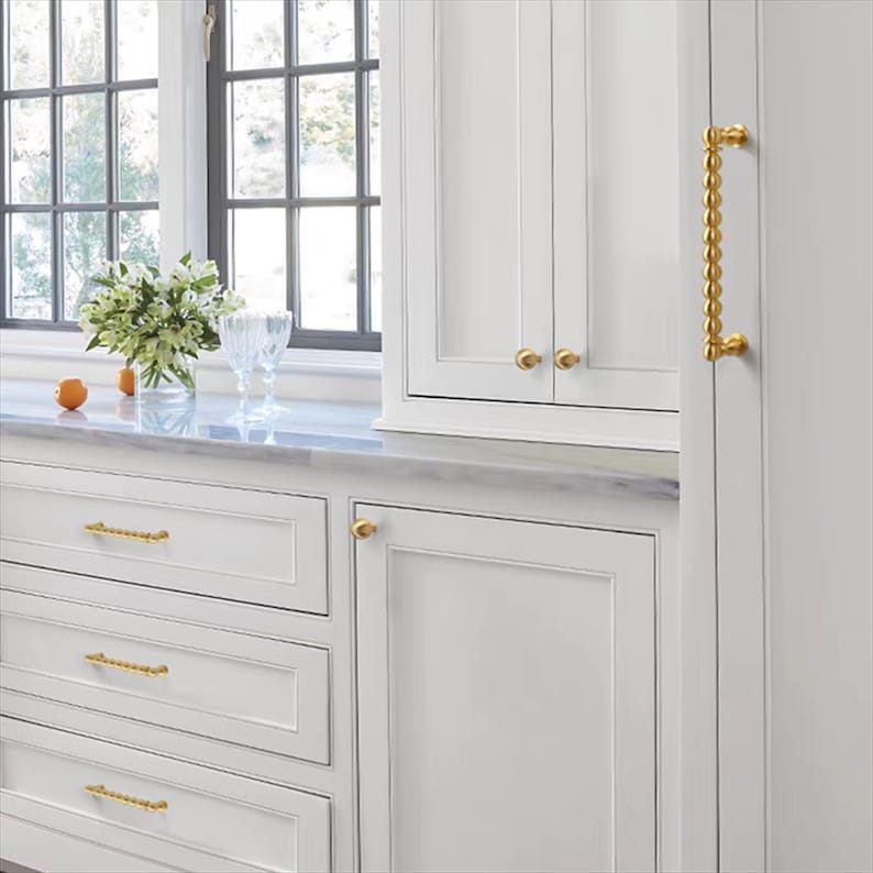 K&ouml;nnte beinhalten: Wei&szlig;e K&uuml;chenschr&auml;nke mit goldfarbener Hardware. Die Schr&auml;nke haben ein klares, modernes Design mit einer Marmorarbeitsplatte. Die goldfarbene Hardware umfasst Kn&ouml;pfe und einen langen Griff. Ein Fenster mit Aussicht befindet sich im Hintergrund.