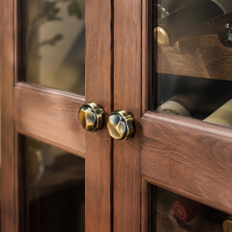 May include: Close-up of a wooden cabinet with glass doors. The cabinet has a dark brown finish and is fitted with two antique brass knobs. The glass doors reveal a glimpse of wine bottles inside, suggesting a wine storage cabinet.