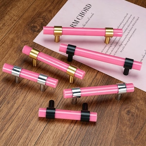 Può includere: Una collezione di sei maniglie per armadietti cilindriche rosa con staffe di montaggio metalliche in finiture oro, argento e nero. Ogni maniglia è composta da una barra rosa liscia fissata da due supporti metallici abbinati.