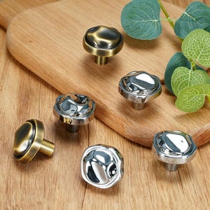 May include: A selection of cabinet knobs in antique brass and polished chrome. The knobs have an unusual, irregular shape and are mounted on short stems. The knobs are displayed on a wooden surface.