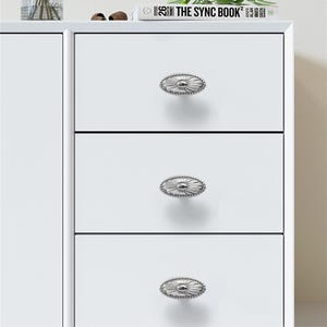 K&ouml;nnte beinhalten: Ein wei&szlig;er Schrank mit drei Schubladen, jede mit einem ovalen, silberfarbenen Knopf mit dekorativem Rand. Die Kn&ouml;pfe sind mittig auf den Schubladen angebracht. Oben auf dem Schrank liegen ein Buch und eine Vase.