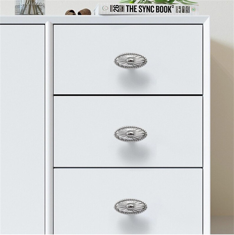 K&ouml;nnte beinhalten: Ein wei&szlig;er Schrank mit drei Schubladen, jede mit einem ovalen, silberfarbenen Knopf mit dekorativem Rand. Die Kn&ouml;pfe sind mittig auf den Schubladen angebracht. Oben auf dem Schrank liegen ein Buch und eine Vase.