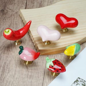 Pode incluir: Uma cole&ccedil;&atilde;o de puxadores de arm&aacute;rio de cer&acirc;mica em v&aacute;rias formas de frutas e vegetais. Os puxadores incluem uma pimenta vermelha, um cora&ccedil;&atilde;o rosa, um cora&ccedil;&atilde;o vermelho, uma pimenta rosa, uma espiga de milho e um morango. Cada puxador tem uma base de lat&atilde;o.