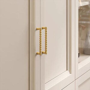 K&ouml;nnte beinhalten: Nahaufnahme einer Schrankt&uuml;r mit zwei goldfarbenen, perlenbesetzten Griffen. Die Schrankt&uuml;r ist cremefarben und die Griffe sind vertikal ausgerichtet. Die Griffe haben ein dekoratives, elegantes Design.