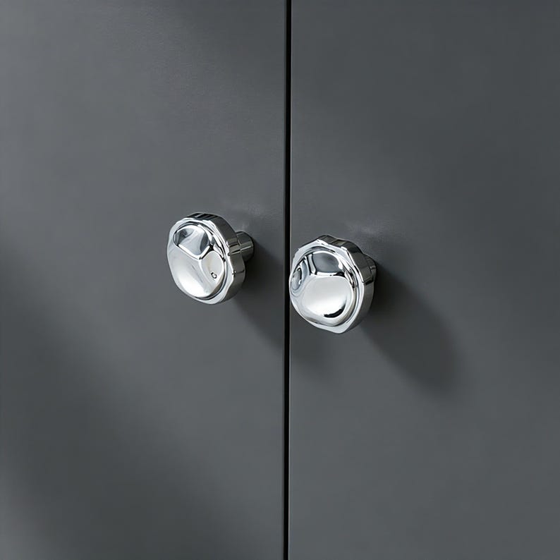 May include: Two polished chrome cabinet knobs with a distinctive, faceted design. The knobs are mounted on a dark grey cabinet door, creating a modern and elegant aesthetic. The knobs are round with a flower-like shape.