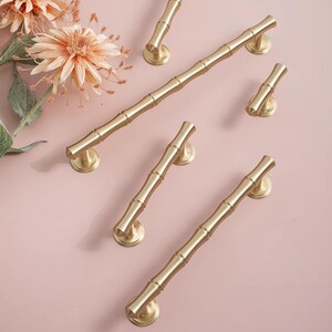 May include: A selection of gold-coloured bamboo-style cabinet pulls and handles. The hardware is arranged on a light pink surface, with decorative flowers in the upper left corner. The pulls vary in size and shape.