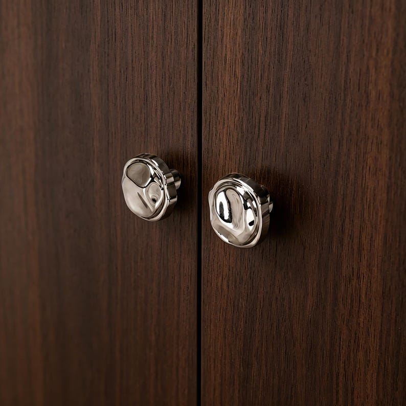 May include: Two polished silver cabinet knobs with an unusual, organic design. They are mounted on a dark brown wooden cabinet door, highlighting the contrast between the metal and the wood grain.