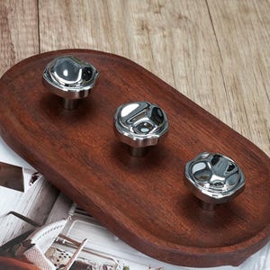 May include: Three polished chrome cabinet knobs with a distinctive, textured surface. They are displayed on a dark brown, oval wooden tray, offering a contemporary and stylish look. The knobs are approximately 3.8 cm in diameter.