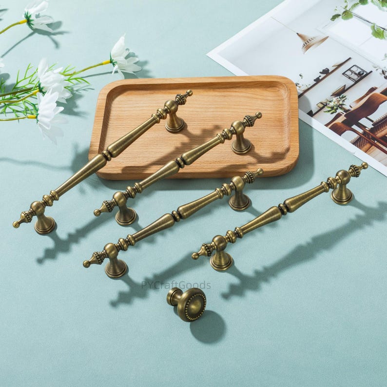 Peut inclure: Ensemble de quincaillerie de meubles en laiton antique, comprenant plusieurs poign&eacute;es et un bouton. Les poign&eacute;es, de diff&eacute;rentes tailles, pr&eacute;sentent des d&eacute;tails orn&eacute;s. L'ensemble est pr&eacute;sent&eacute; sur un plateau en bois.