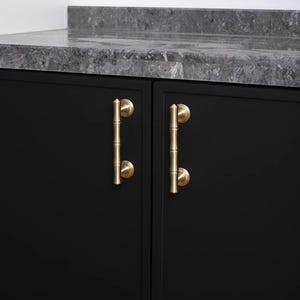 May include: A pair of gold bamboo-style handles on a black cabinet. The handles are mounted vertically on the cabinet doors. A grey countertop is visible above the cabinet. The handles are made of metal.