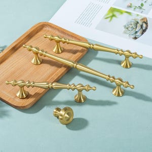 Peut inclure: Ensemble de quincaillerie de meubles dor&eacute;e, comprenant plusieurs poign&eacute;es et un bouton. Les poign&eacute;es pr&eacute;sentent des d&eacute;tails d&eacute;coratifs et des formes vari&eacute;es. Pr&eacute;sent&eacute; sur un plateau en bois et une surface bleu clair.