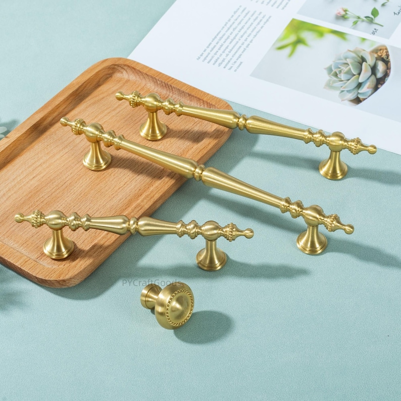 Peut inclure: Ensemble de quincaillerie de meubles dor&eacute;e, comprenant plusieurs poign&eacute;es et un bouton. Les poign&eacute;es pr&eacute;sentent des d&eacute;tails d&eacute;coratifs et des formes vari&eacute;es. Pr&eacute;sent&eacute; sur un plateau en bois et une surface bleu clair.