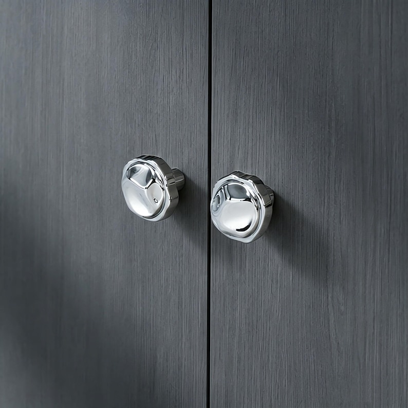 May include: Two polished silver cabinet knobs with a distinctive, textured pattern. They are fixed to a dark grey wooden cabinet door, offering a contemporary and stylish appearance. The knobs have a reflective surface.