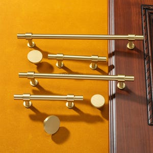 K&ouml;nnte beinhalten: Eine Sammlung goldfarbener Schrankbeschl&auml;ge, darunter Stangengriffe und runde Kn&ouml;pfe. Die Griffe sind unterschiedlich lang, und die Kn&ouml;pfe sind kreisf&ouml;rmig. Die Beschl&auml;ge werden vor einem senfgelben Hintergrund pr&auml;sentiert, mit einem sichtbaren Holzrahmen.