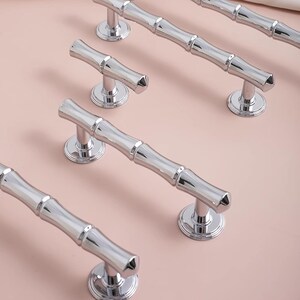 May include: A collection of polished chrome cabinet handles, each with a bamboo-style design, are displayed on a light pink surface. The handles feature a ridged, cylindrical design and are mounted on circular bases.