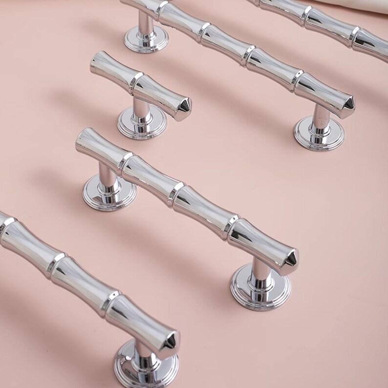 May include: A collection of polished chrome cabinet handles, each with a bamboo-style design, are displayed on a light pink surface. The handles feature a ridged, cylindrical design and are mounted on circular bases.