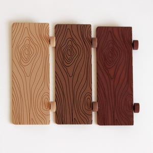 May include: Three wooden rectangular panels in varying shades of brown, beige, and reddish-brown, each with a wood grain pattern. The panels are connected by small, cylindrical, beige and brown hinges, suggesting a shutter or decorative element.