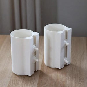 Op de afbeelding: Twee cilindrische witte kunststof houders met een verticaal rechthoekig opzetstuk aan de zijkant. Elk opzetstuk is voorzien van twee kleine uitstekende pinnetjes. De objecten hebben een gladde, matte afwerking en een open bovenkant, rechtopstaand.