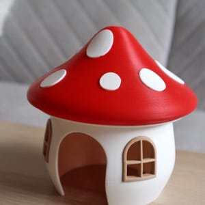 May include: A charming mushroom house featuring a red cap with white spots. The house has a white base with arched openings and brown window frames. This decorative item is a unique home decor piece, likely for a child's room.