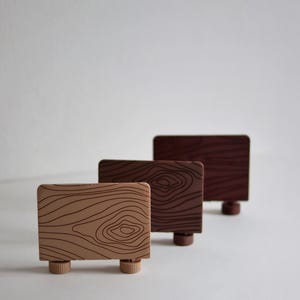 May include: Three wooden toy fences in varying shades of brown. Each fence has a rectangular shape with a wood grain pattern and is supported by two small, round feet. The fences are arranged in a row, with the lightest shade in front.