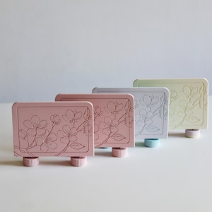 May include: Four rectangular, pastel-colored bottle stoppers. Each stopper features a floral design and rests on a small, cylindrical base. The colors include pink, light pink, gray, and pale green.