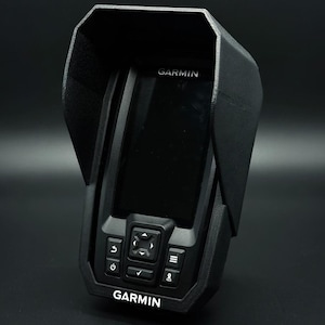 Puede incluir: Un buscador de peces Garmin negro con una cubierta protectora. El dispositivo tiene una pantalla, botones y el logotipo de Garmin en blanco. La cubierta tiene una superficie texturizada y una extensi&oacute;n tipo visera.