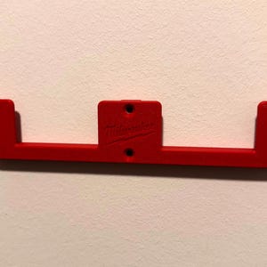 May include: A bright red, plastic bracket with a rectangular center section and two L-shaped end pieces. The bracket has several holes for mounting and the Milwaukee logo is visible. The bracket is designed to be mounted on a wall.