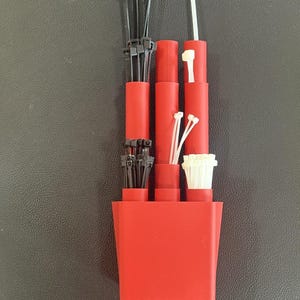 Puede incluir: Un organizador de plástico rojo con varias bridas y otros pequeños componentes blancos y negros. El organizador tiene varios compartimentos cilíndricos que contienen paquetes de bridas negras, bridas blancas y otros pequeños componentes blancos.