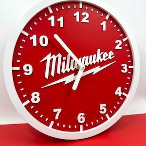 May include: A round wall clock with a red face and white numerals and hands. The clock has a white border and the word "Milwaukee" in white with a lightning bolt design. The clock is a decorative item.