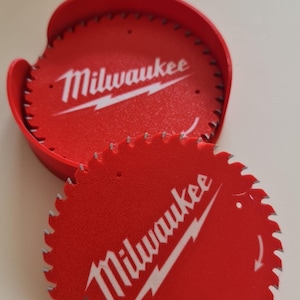 May include: Red circular saw blades with the "Milwaukee" logo in white. One blade is in a red holder. The blades have a toothed edge. The image is taken from a slightly elevated angle.