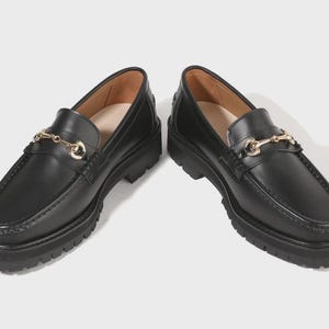 Peut inclure: Une paire de mocassins en cuir noir avec un d&eacute;tail mors en m&eacute;tal dor&eacute; sur le dessus. Ces chaussures pr&eacute;sentent une &eacute;paisse semelle crant&eacute;e en caoutchouc noir avec un motif antid&eacute;rapant et un bout arrondi, offrant une finition polie.