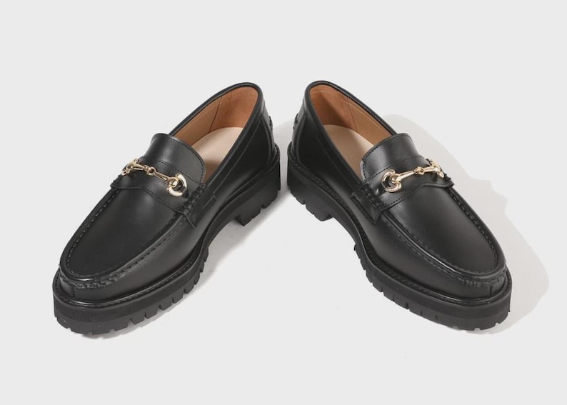 Peut inclure: Une paire de mocassins en cuir noir avec un d&eacute;tail mors en m&eacute;tal dor&eacute; sur le dessus. Ces chaussures pr&eacute;sentent une &eacute;paisse semelle crant&eacute;e en caoutchouc noir avec un motif antid&eacute;rapant et un bout arrondi, offrant une finition polie.
