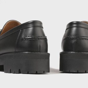 Peut inclure: Une paire de mocassins en cuir noir dot&eacute;s d'une semelle &eacute;paisse et crant&eacute;e avec des rainures profondes. Les chaussures pr&eacute;sentent une finition lisse, un bout arrondi et des coutures apparentes le long du contrefort et du bord sup&eacute;rieur.