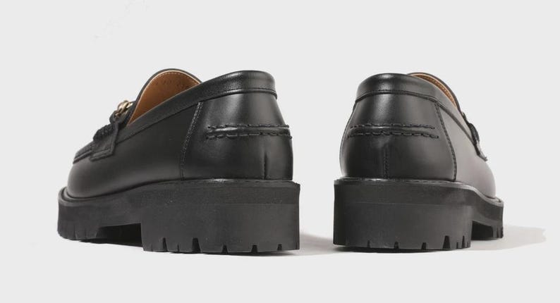 Peut inclure: Une paire de mocassins en cuir noir dot&eacute;s d'une semelle &eacute;paisse et crant&eacute;e avec des rainures profondes. Les chaussures pr&eacute;sentent une finition lisse, un bout arrondi et des coutures apparentes le long du contrefort et du bord sup&eacute;rieur.