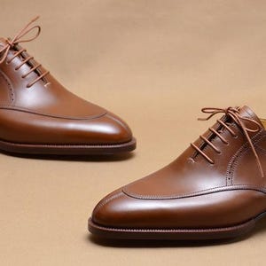 May include: A pair of polished brown leather oxford shoes featuring a split-toe design, thin brown laces, and a stacked wooden heel. Each shoe includes a light-colored wooden shoe tree inserted inside to maintain the shape of the footwear.