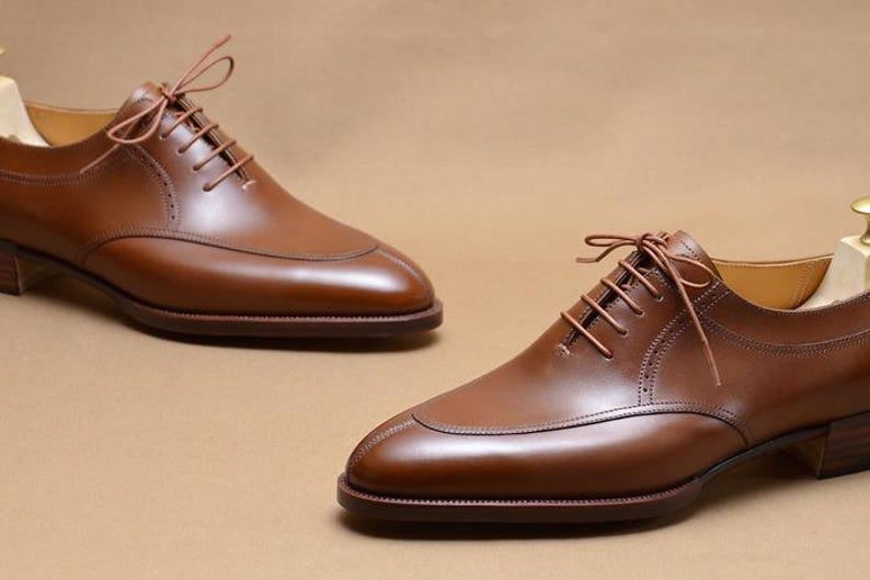 May include: A pair of polished brown leather oxford shoes featuring a split-toe design, thin brown laces, and a stacked wooden heel. Each shoe includes a light-colored wooden shoe tree inserted inside to maintain the shape of the footwear.