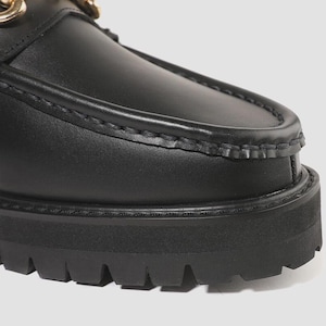 Peut inclure: Un mocassin en cuir noir avec une couture apparente sur le dessus, une semelle &eacute;paisse en caoutchouc crant&eacute; et un d&eacute;tail m&eacute;tallique dor&eacute; en forme de mors. La chaussure pr&eacute;sente une finition lisse et une semelle ext&eacute;rieure &agrave; crampons.