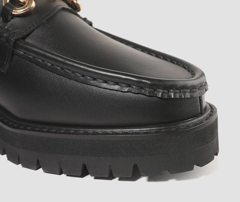 Peut inclure: Un mocassin en cuir noir avec une couture apparente sur le dessus, une semelle &eacute;paisse en caoutchouc crant&eacute; et un d&eacute;tail m&eacute;tallique dor&eacute; en forme de mors. La chaussure pr&eacute;sente une finition lisse et une semelle ext&eacute;rieure &agrave; crampons.