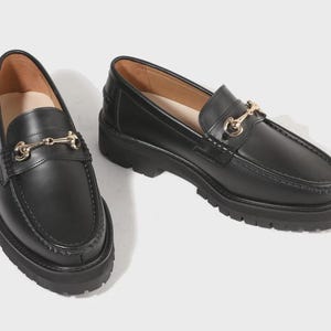 Peut inclure: Une paire de mocassins en cuir noir avec un d&eacute;tail mors en m&eacute;tal dor&eacute; sur le dessus. Ces chaussures pr&eacute;sentent un bout arrondi, une couture en relief sur l'avant et une &eacute;paisse semelle crant&eacute;e en caoutchouc noir pour plus de hauteur.