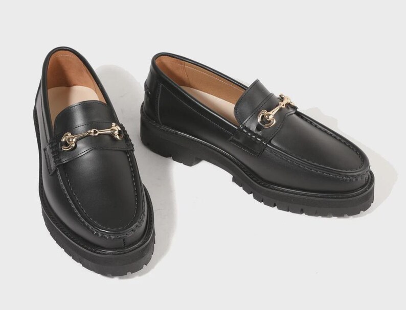 Peut inclure: Une paire de mocassins en cuir noir avec un d&eacute;tail mors en m&eacute;tal dor&eacute; sur le dessus. Ces chaussures pr&eacute;sentent un bout arrondi, une couture en relief sur l'avant et une &eacute;paisse semelle crant&eacute;e en caoutchouc noir pour plus de hauteur.