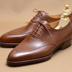 May include: A pair of brown leather oxford shoes with a split-toe design, thin brown laces, and a stacked wooden heel. Each shoe features a light-colored wooden shoe tree inserted inside to maintain the shape of the leather upper.