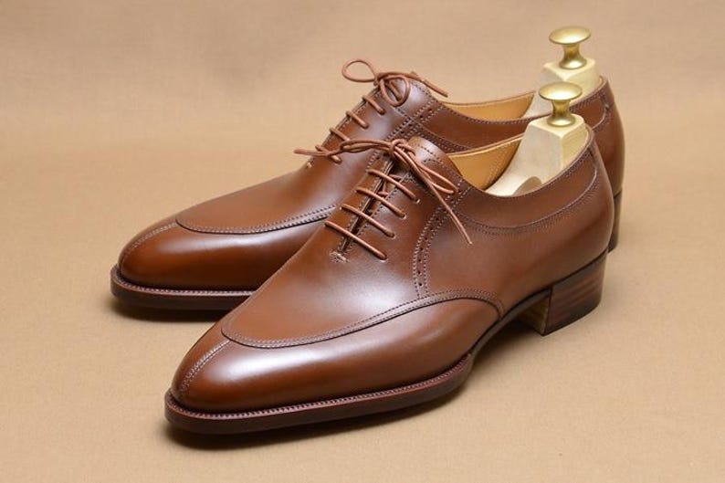 May include: A pair of brown leather oxford shoes with a split-toe design, thin brown laces, and a stacked wooden heel. Each shoe features a light-colored wooden shoe tree inserted inside to maintain the shape of the leather upper.