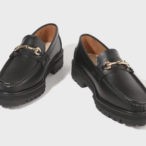 Peut inclure: Une paire de mocassins en cuir noir avec un d&eacute;tail de mors en m&eacute;tal dor&eacute; sur le dessus. Ces chaussures pr&eacute;sentent une &eacute;paisse semelle crant&eacute;e noire avec un motif de bande de roulement profond et un bout arrondi, offrant une base solide.
