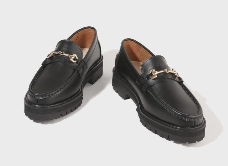 Peut inclure: Une paire de mocassins en cuir noir avec un d&eacute;tail de mors en m&eacute;tal dor&eacute; sur le dessus. Ces chaussures pr&eacute;sentent une &eacute;paisse semelle crant&eacute;e noire avec un motif de bande de roulement profond et un bout arrondi, offrant une base solide.