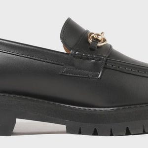 Peut inclure: Un mocassin en cuir noir dot&eacute; d'une &eacute;paisse semelle en caoutchouc crant&eacute;e et d'un d&eacute;tail mors en m&eacute;tal dor&eacute; sur le dessus. La chaussure pr&eacute;sente une couture en relief sur le bout, un contrefort structur&eacute; et une finition lisse et brillante.