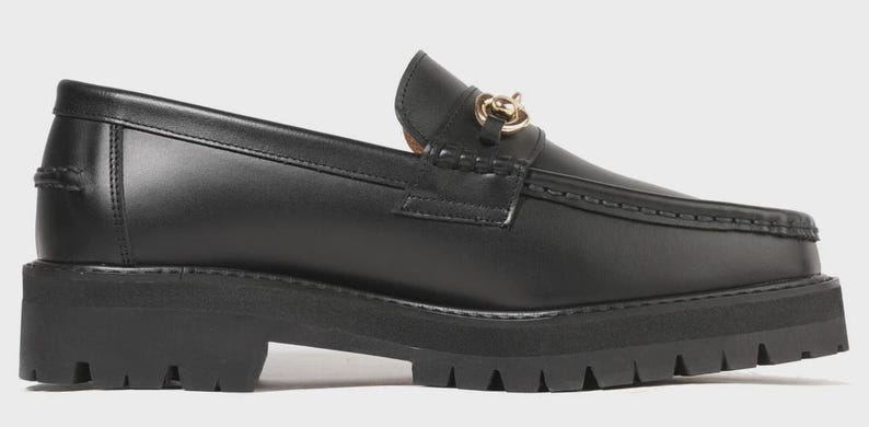Peut inclure: Un mocassin en cuir noir dot&eacute; d'une &eacute;paisse semelle en caoutchouc crant&eacute;e et d'un d&eacute;tail mors en m&eacute;tal dor&eacute; sur le dessus. La chaussure pr&eacute;sente une couture en relief sur le bout, un contrefort structur&eacute; et une finition lisse et brillante.