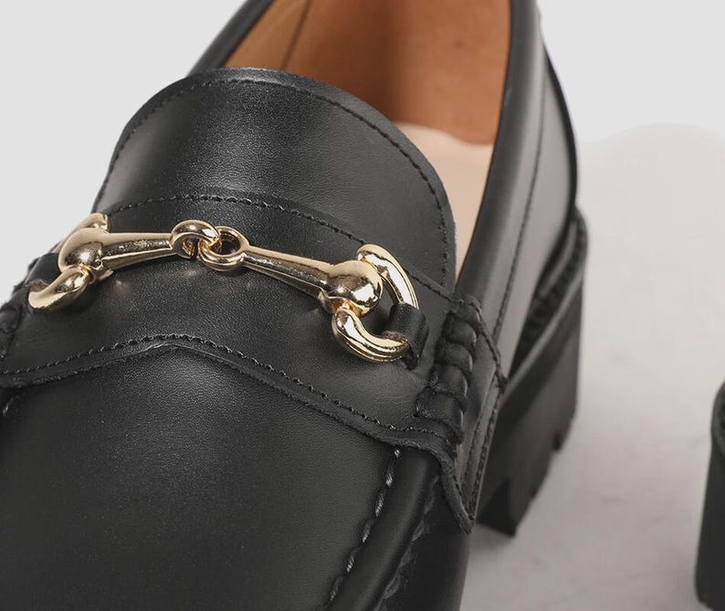 Peut inclure: Un mocassin en cuir noir dot&eacute; d'un mors en m&eacute;tal dor&eacute; brillant sur le dessus. La chaussure pr&eacute;sente une tige en cuir lisse avec des coutures apparentes le long de l'empeigne et une semelle noire &eacute;paisse et crant&eacute;e pour un profil robuste.