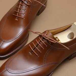 May include: A pair of polished brown leather oxford shoes with a split-toe design and matching brown laces. One shoe features a light-colored wooden shoe tree with a brass-colored knob inserted inside to maintain its shape and structure.